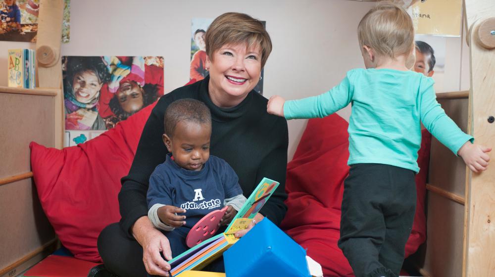 A woman reads to one child on her lap while another walks nearby.