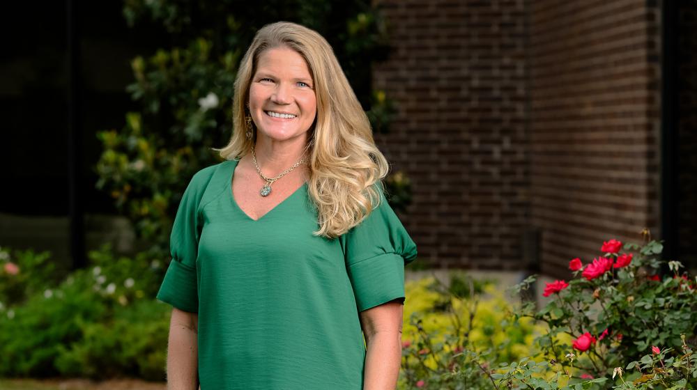 A smiling person wearing green and standing outside in front of roses.