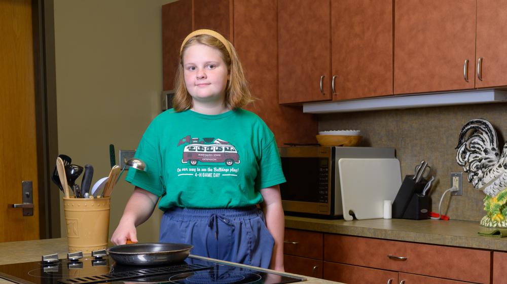A smiling child wearing a green shirt and standing in a kitchen.