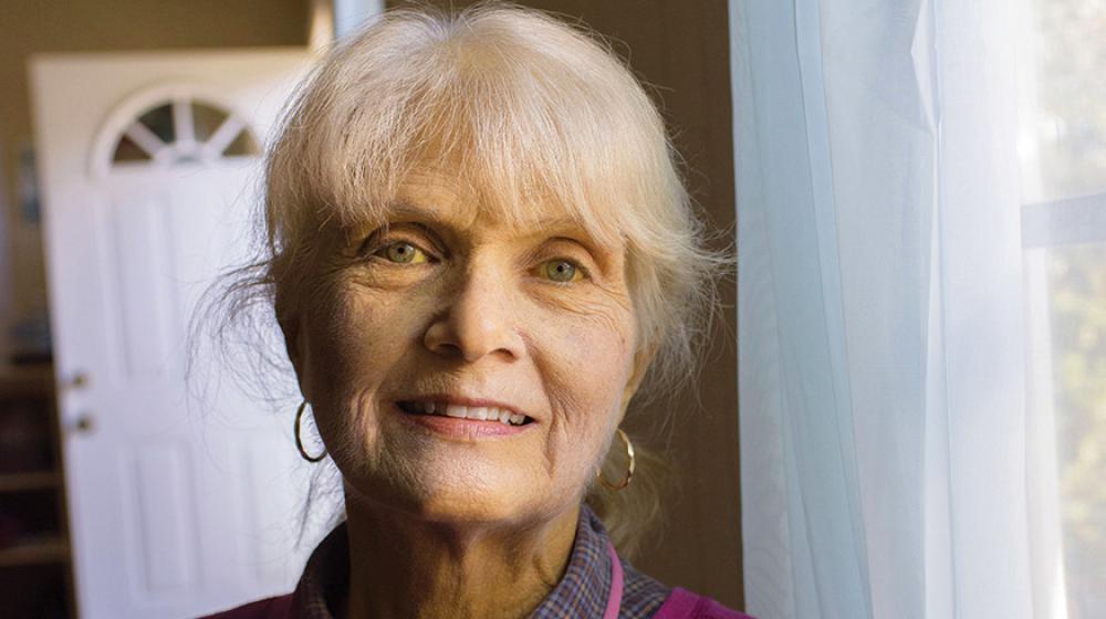 An smiling elderly woman stands next to a window. 