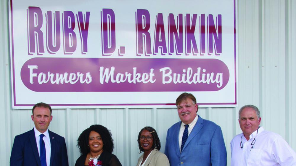Five people, three men and two women, stand in front of a Farmers Market Building sign