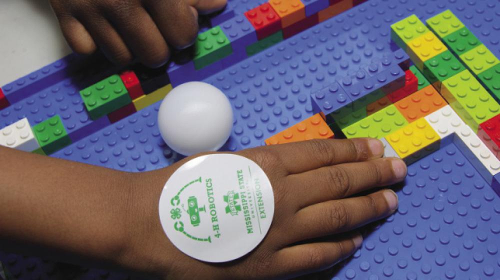 Hand of a child with a 4-H sticker placing colorful LEGO bricks for 4-H Robotics. A child uses colorful LEGO bricks at a 4-H Robotics competition.