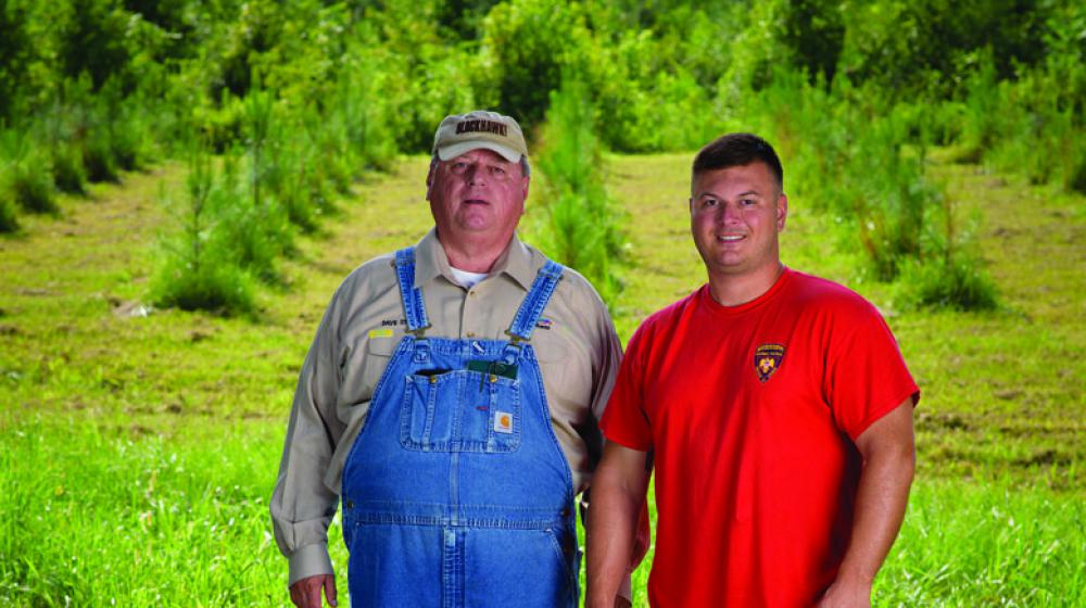 •	A man in overalls stands beside a man in a red T-shirt in front of a tree farm