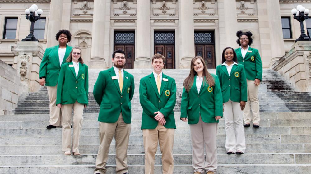 Seven teenagers wearing green blazers and tan slacks form a U standing on steps in front of six gray columns.