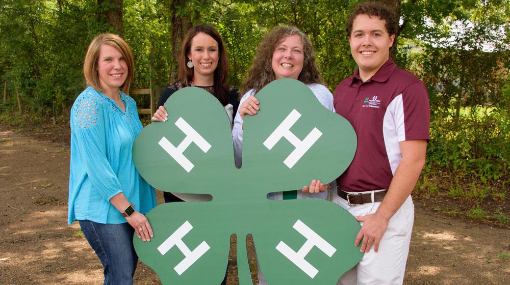 Three women and one man hold a large 4-H clover