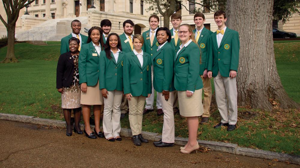 A group of high school girls and boys wearing khaki bottoms and green 4-H blazers stand in a group in front of the state capitol building.