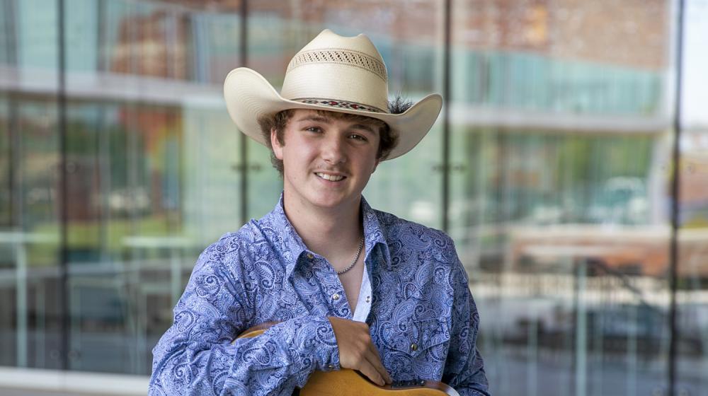 A young man wearing a cowboy hat, paisley collared shirt, and a belt buckle on jeans, holding a guitar and smiling.