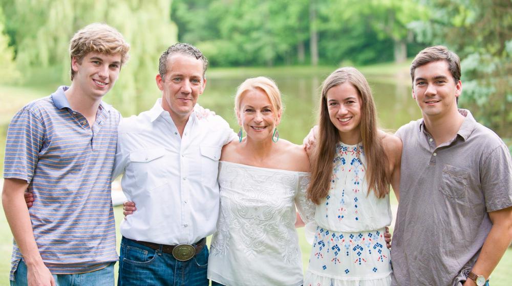 Family photo of 2 parents and 3 young adults standing with arms around each other in front of a small body of water.
