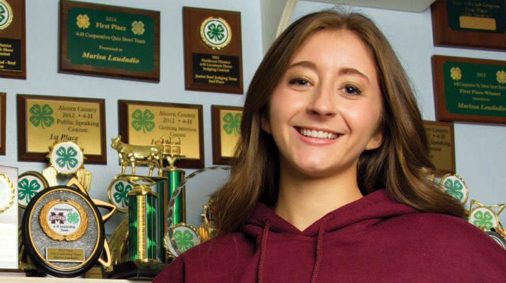 A girl wearing a maroon sweatshirt stands in front of a trophy display with her hands on her hips.