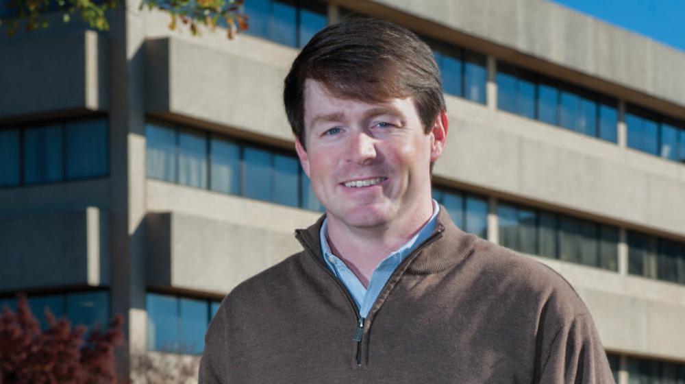 A man with hands in his pockets stands smiling in front of a large concrete building.