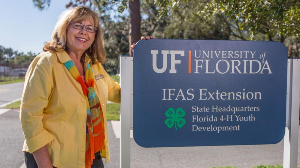 A blonde woman with glasses, wearing a yellow shirt and a motley scarf, stands smiling on a sidewalk in front of trees beside a sign marking “UF University IFAS Extension State Headquarters Florida 4-H Youth Development.”
