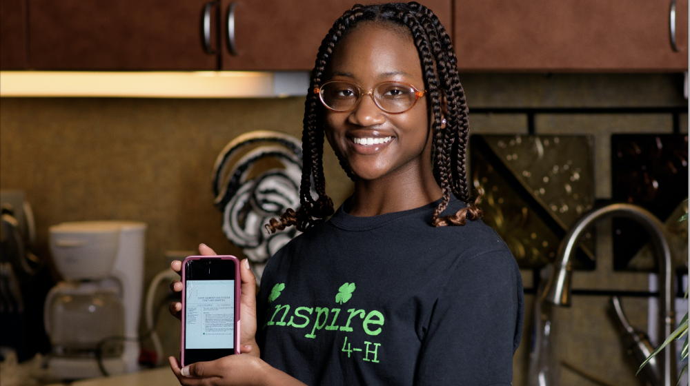 A smiling teenage girl stands behind a table covered in ingredients for a recipe displayed on a phone screen she holds in front of her. 