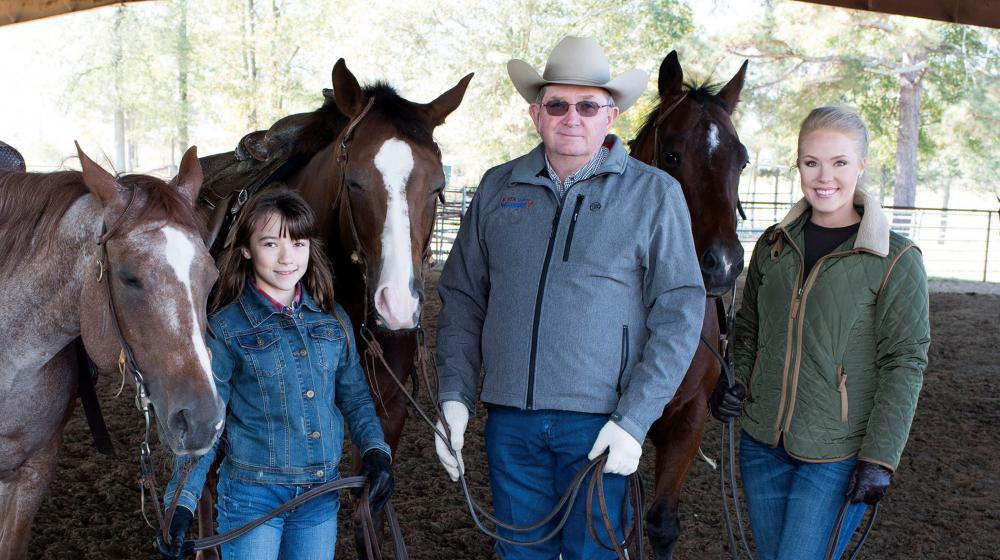 A man and two girls stand in a barn with three horses.