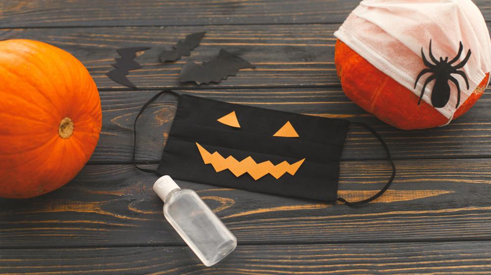 A black jack-o-lantern mask, bottle of sanitizer, and two pumpkins on a wooden backdrop.