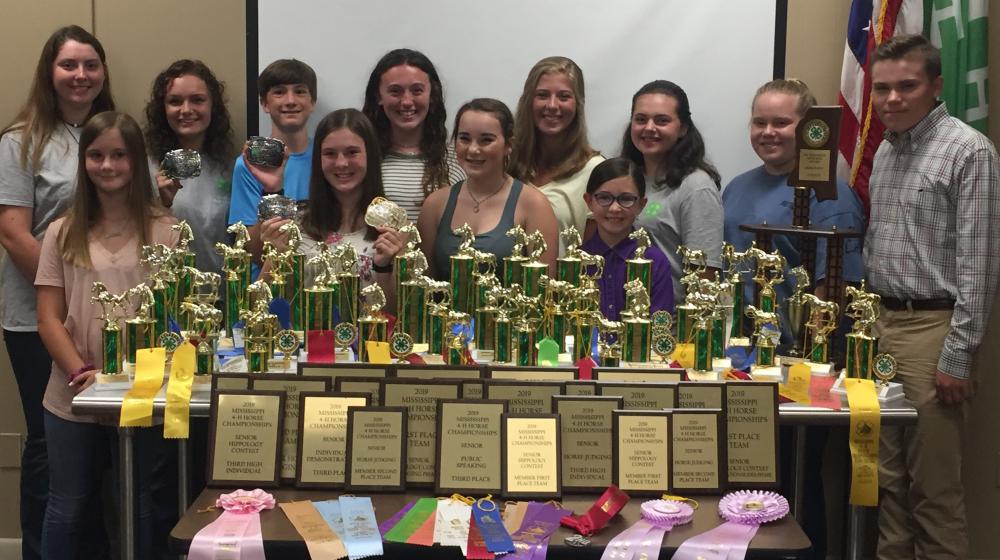 Twelve people stand behind a table covered in trophies, plaques, and different colored ribbons.