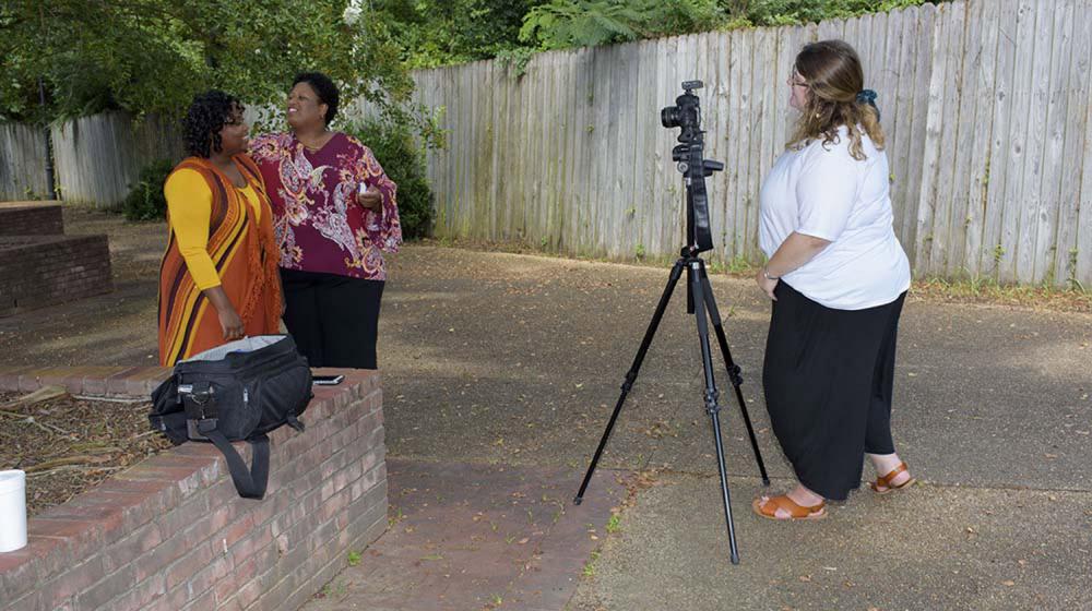 A woman wearing a white shirt stands behind a camera as two women in front of the camera smile.