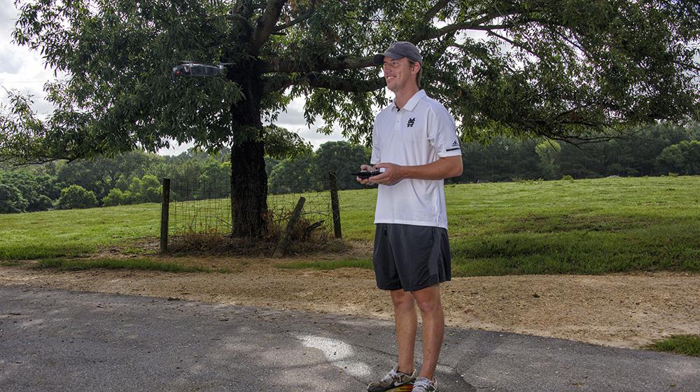 A man wearing a white shirt and blue baseball cap smiles as he looks at a drone flying through the air.