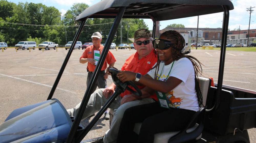 A young girl wearing goggles over her eyes sits next to an older man in a golf cart. 