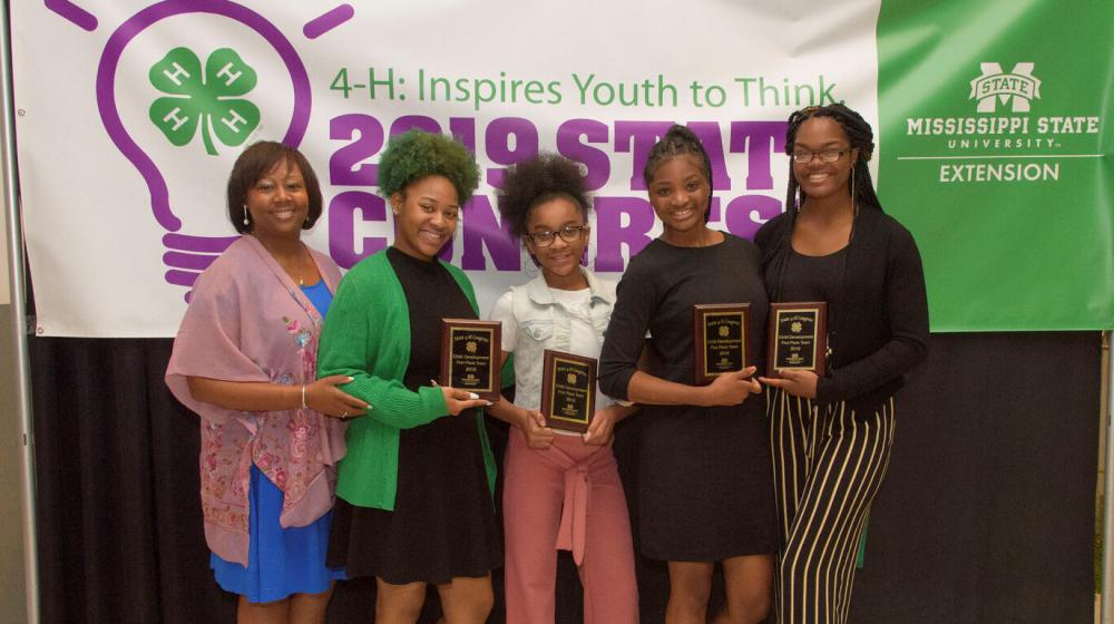 Four teenage girls hlding plaques and an older woman stand in front of a purple, white, and green 4-H poster.