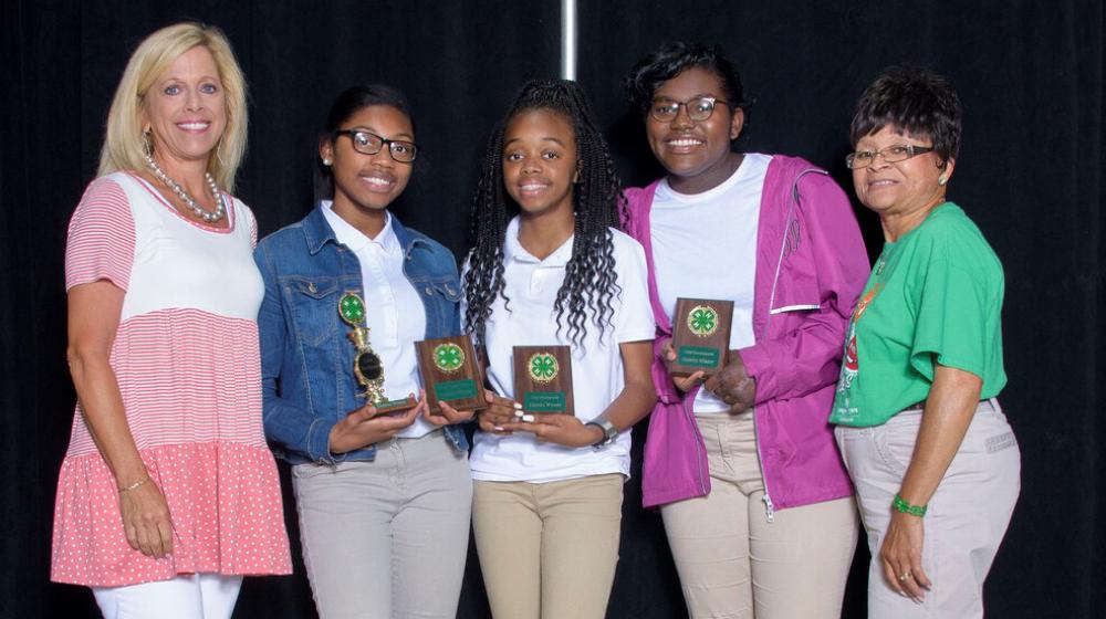 Three young girls holding 4-H plaques stand between two older women.