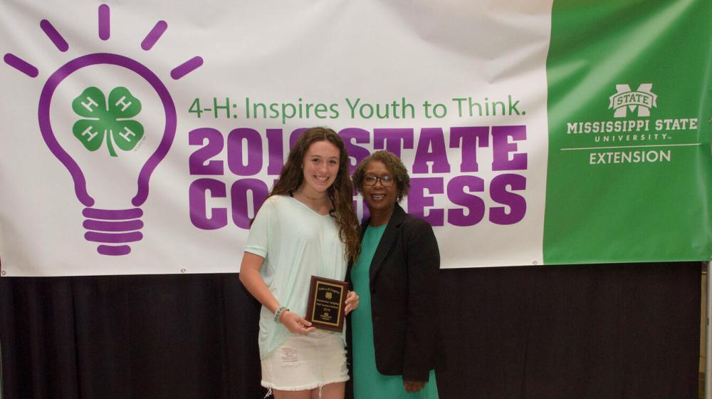A teenage girl holding a plaque stands next to an older woman in front of a purple, green, and white 4-H poster.
