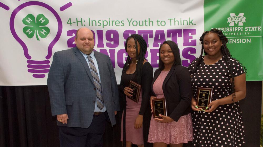 Three teenage girls holding plaques stand next to a man in front of a purple, green, and white 4-H poster.