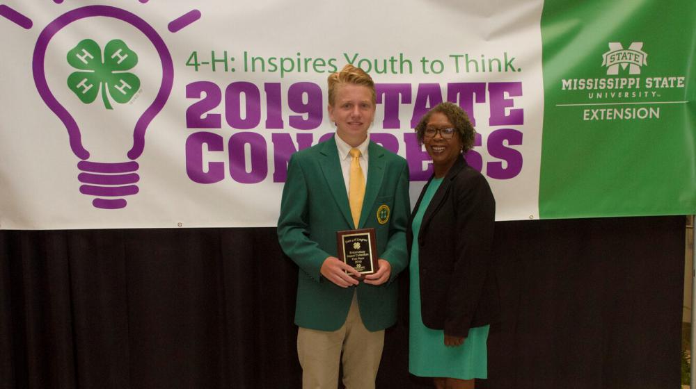 A teenage boy wearing a green blazer and holding a plaque stands next to an older woman in front of a purple, green, and white 4-H poster.