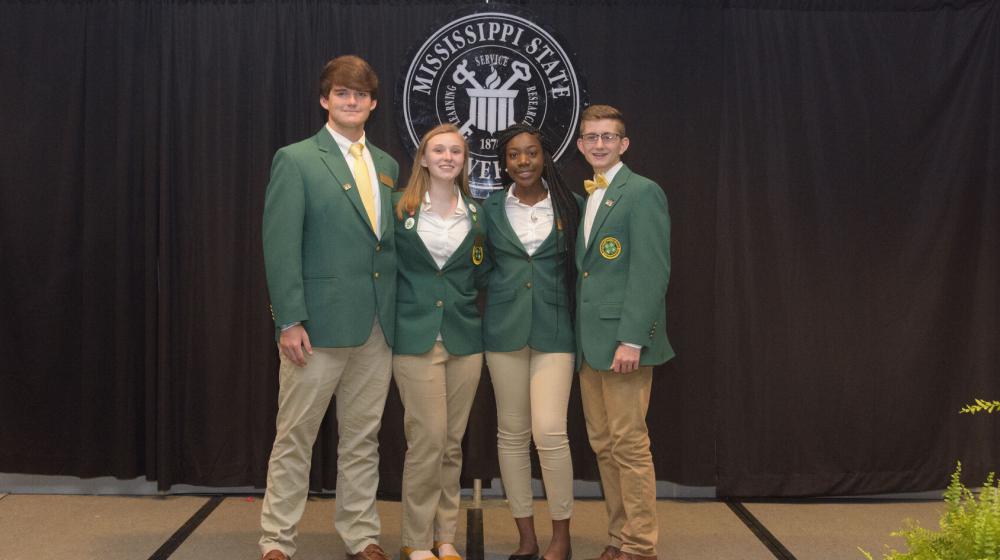 Four teenagers wearing green blazers stand in front of a black curtain.