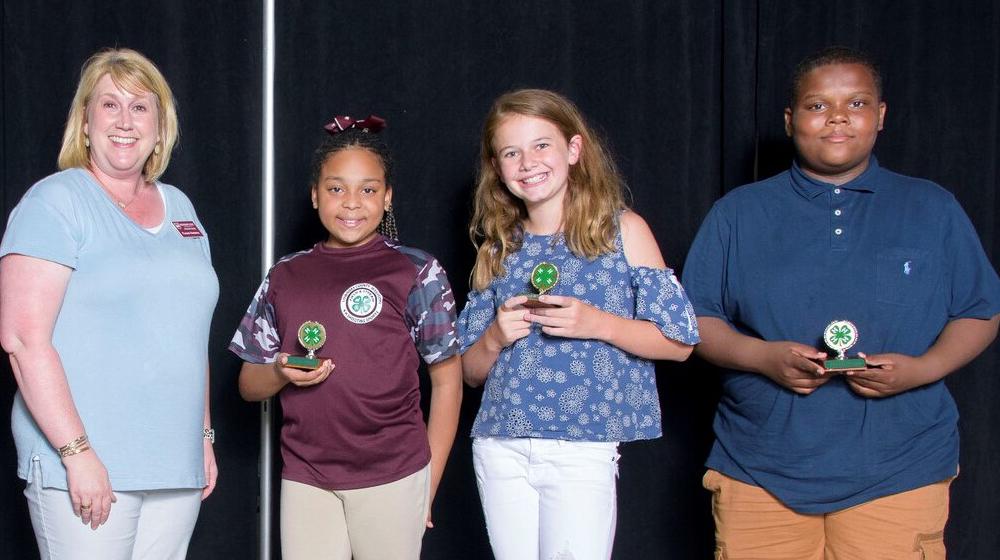 Three young kids holding small green 4-H trophies smile next to an older woman wearing a maroon name tag.