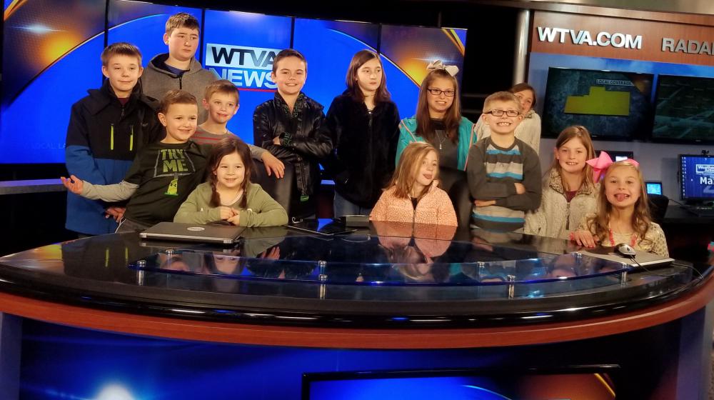 Thirteen young kids stand behind a table with "WTVA News" on the screens in front and behind them. 