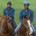 Two boys sit on two horses facing the camera. Both boys are wearing blue shirts and safety helmets.