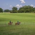 A large, green, grassy field stretches out behind two young men riding brown horses along a fence.