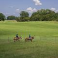 A large, green, grassy field stretches out behind two young men riding brown horses along a fence.