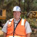 A young man with a metal construction hat and bright orange vest stands in front of his work site with his hands tucked into his blue jeans.