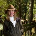 A young woman with brown hair wearing a tan hat with a green jacket and khaki pants leans against a wooden railing.