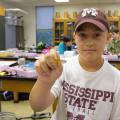 A young boy wearing a Mississippi State t-shirt holds a butterfly-like insect out toward the camera.