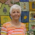 An elderly woman wearing an orange striped shirt stands in front of a large, multicolored, needlepoint county map of Mississippi.