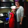 A little girl in a white chef’s hat stands beside her mother