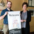 A man in a blue shirt and a woman in a grey shirt with a black cardigan stand holding a banner that says “Welcome…the Square Kosciusko.”