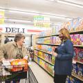 A woman wearing a Mississippi State University mask happily speaks with a woman pushing a grocery cart down an aisle of cereal in a grocery store.