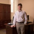 A man wearing a colorful plaid shirt stands in front of a desk.