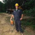 A man wearing a yellow hard hat and holding a chain saw stands in front of a trailer holding harvested timber.