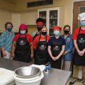 Five young adults and two women wearing black aprons and face masks smile for a group photo in a kitchen.