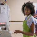 A smiling young girl wearing a green apron holding a measuring cup full of flour.