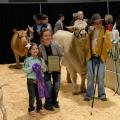 A woman crouches, holding a plaque, with a girl on her left and a boy and steer on her right.