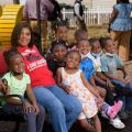 A woman, wearing a red T-shirt listing Rosemary’s Daycare on it, smiles while sitting on a bench with many small children sitting beside her, also smiling.