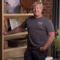 A blonde man wearing a T-shirt with Shuden Farms listed on it standing in front of a bookshelf, smiling.