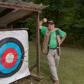 A smiling man wearing a vest and green shirt, standing beside an archery target with three arrows near the bullseye.
