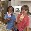 A girl and boy stand in a kitchen and hold up the popsicles they are eating.