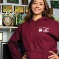 A girl wearing a maroon sweatshirt stands in front of a trophy display with her hands on her hips.